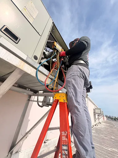 Technician on a ladder servicing rooftop HVAC equipment with gauges
